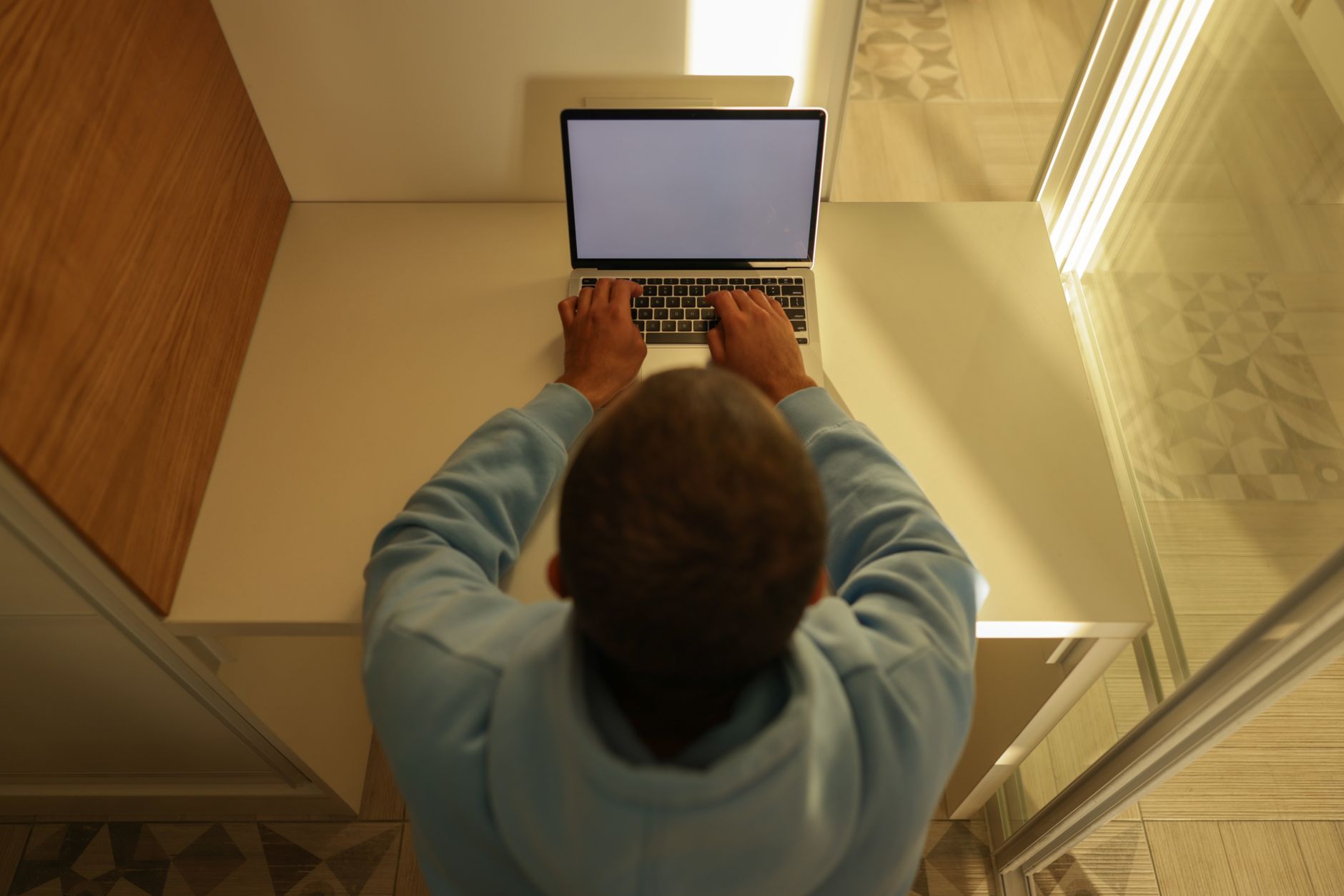 Vue de dessus d’un homme en sweat à capuche bleu qui tape sur un ordinateur portable posé sur un bureau clair.