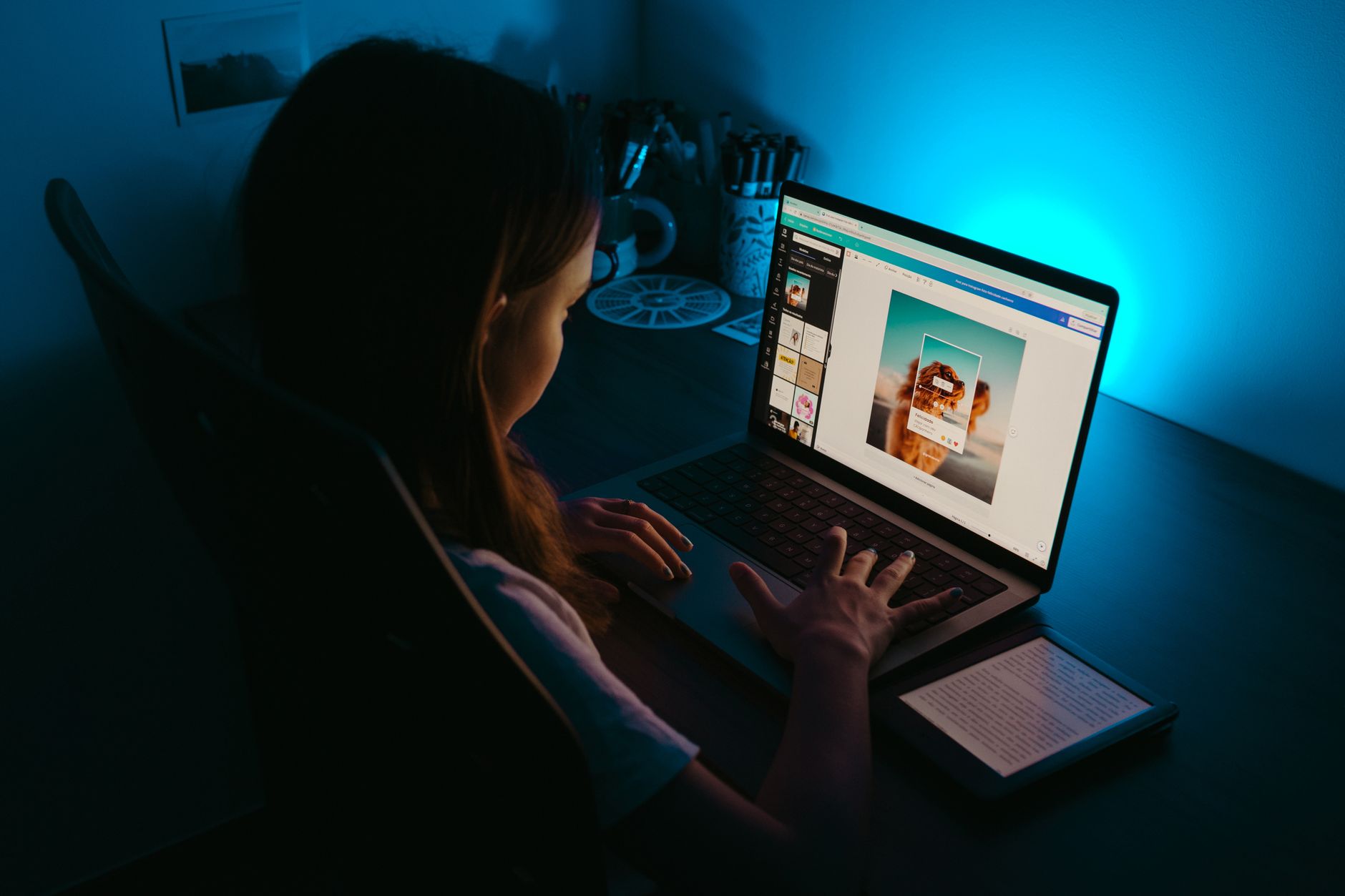 Une personne travaille sur un ordinateur portable à son bureau, dans une pièce sombre éclairée par une lumière bleue.